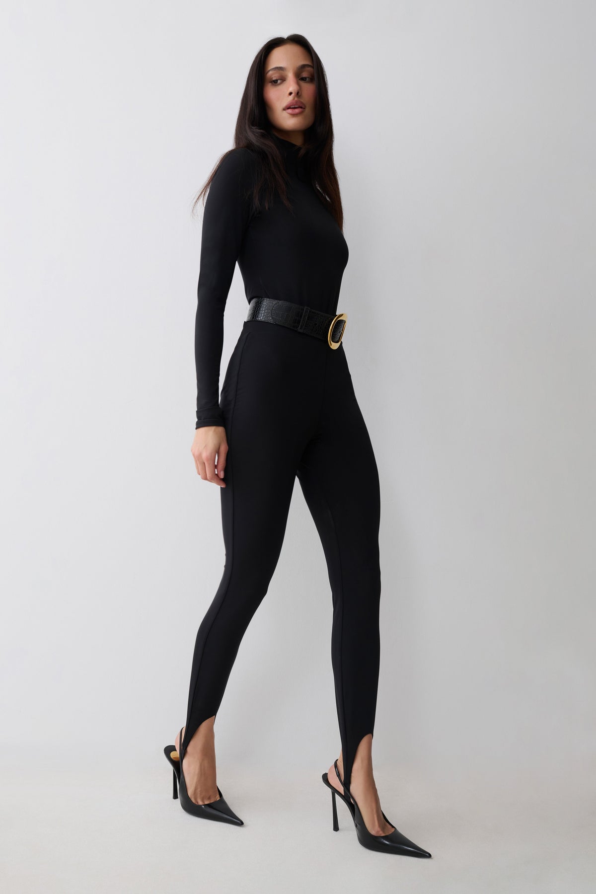 A woman modeling a black long-sleeve bodysuit with black stirrup leggings with a wide black belt featuring a gold oval buckle, paired with black pointed-toe stiletto heels, against a light gray studio background.