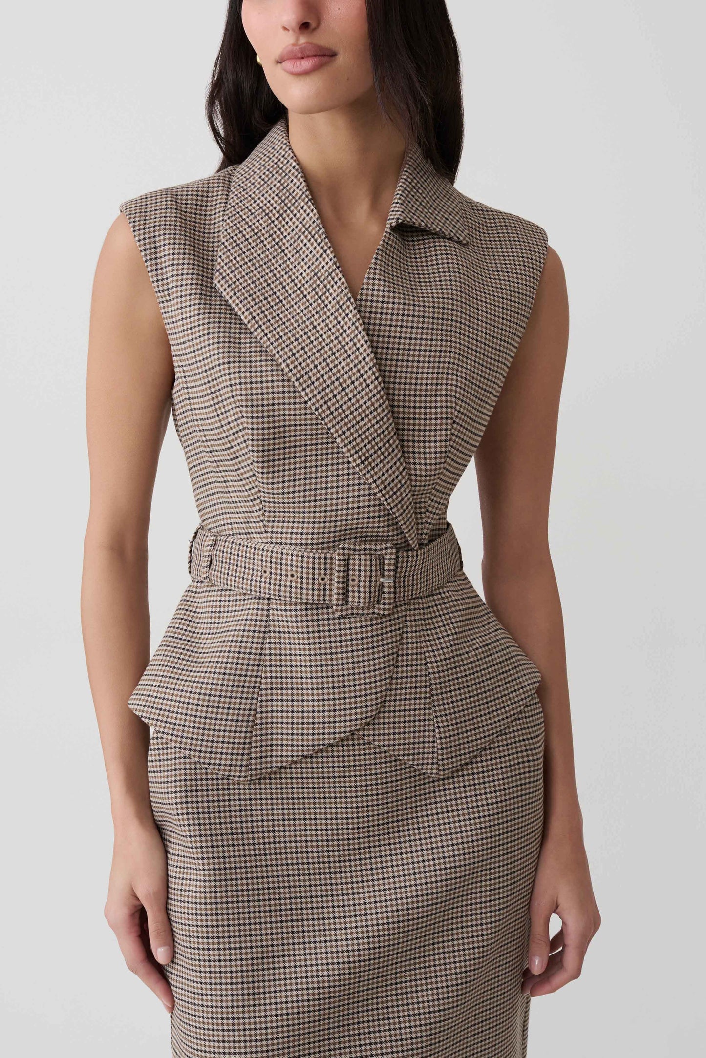 A woman wearing a sleeveless belted wrap dress with a fine brown and beige houndstooth pattern, shown from the neck down against a light gray studio background.