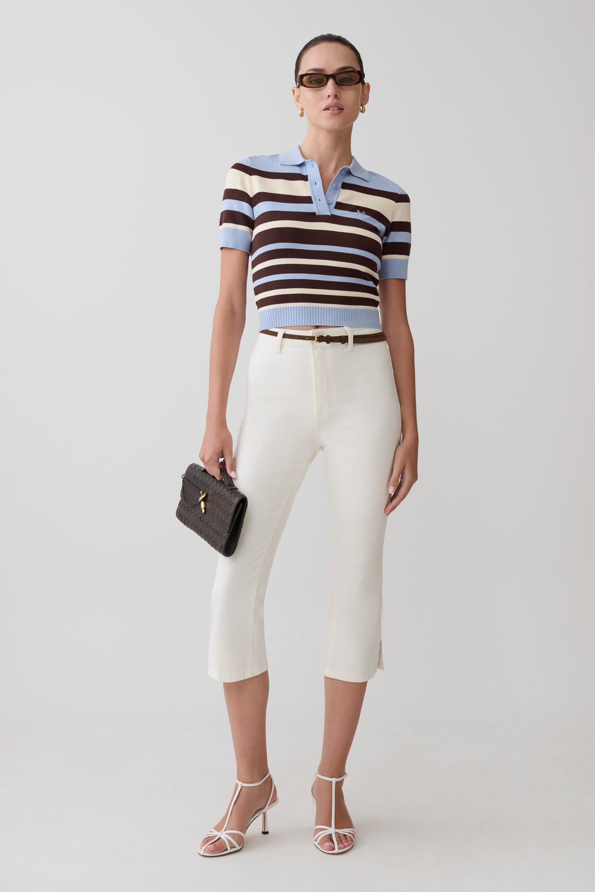 Woman wearing a striped polo shirt and white pants with a black clutch against a white background