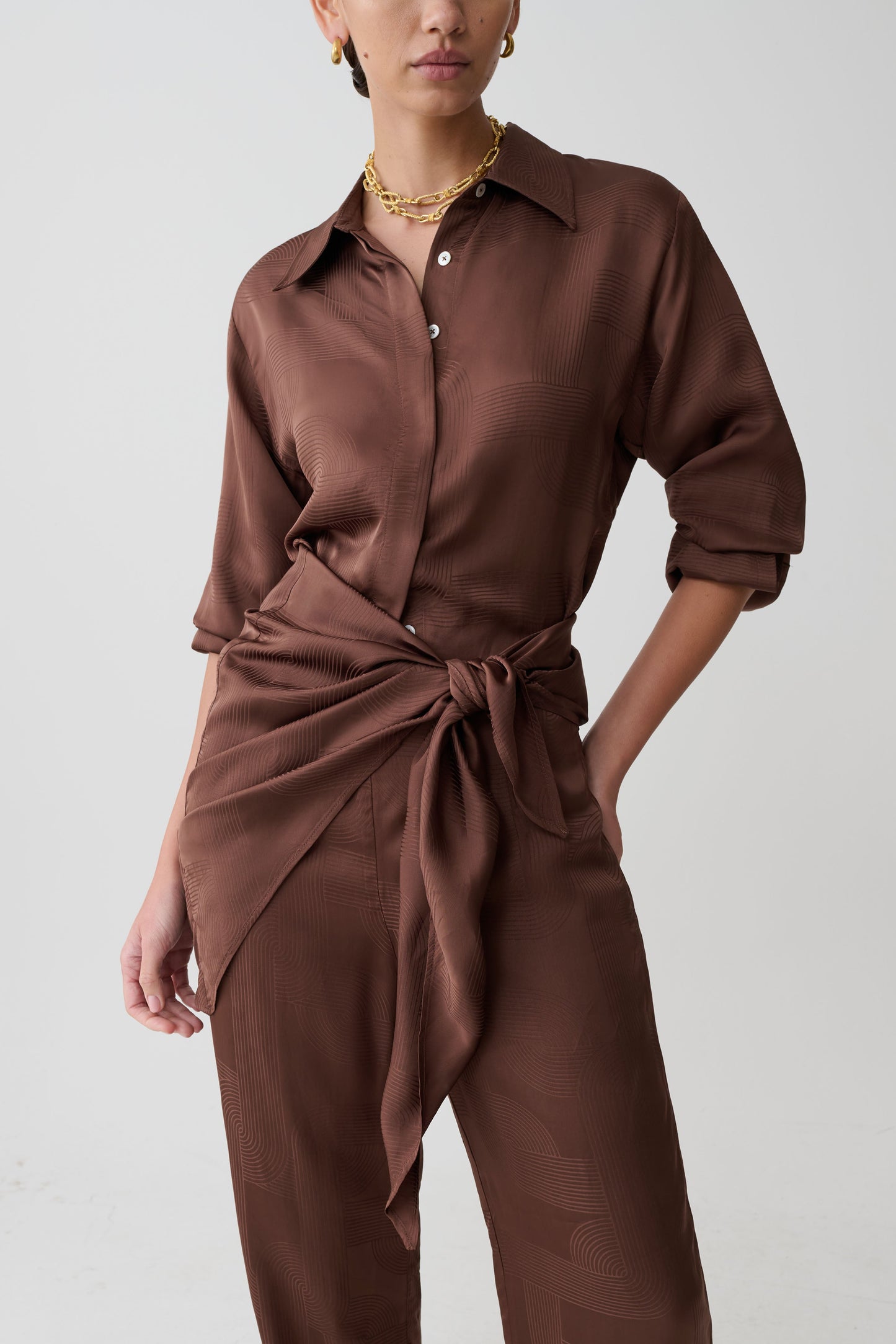 Person wearing a brown shirt and pants with a tied waist on a plain background