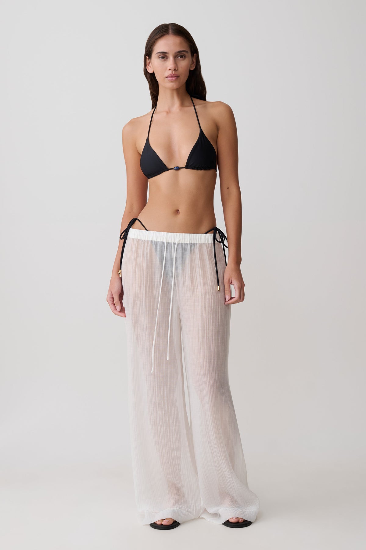 Woman wearing a black bikini top and sheer white drawstring wide pants.