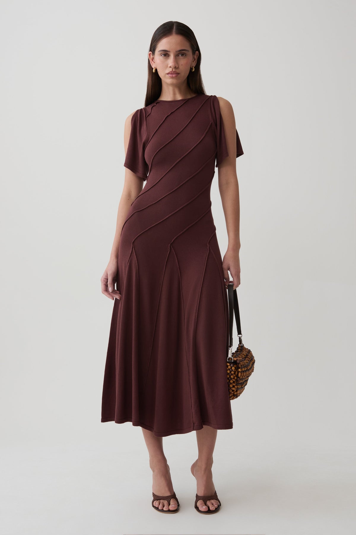 A woman models a dark brown midi dress with draped diagonal paneling and short sleeves with shoulder cutouts, paired with brown strappy heels and a patterned handbag, against a light gray studio background.