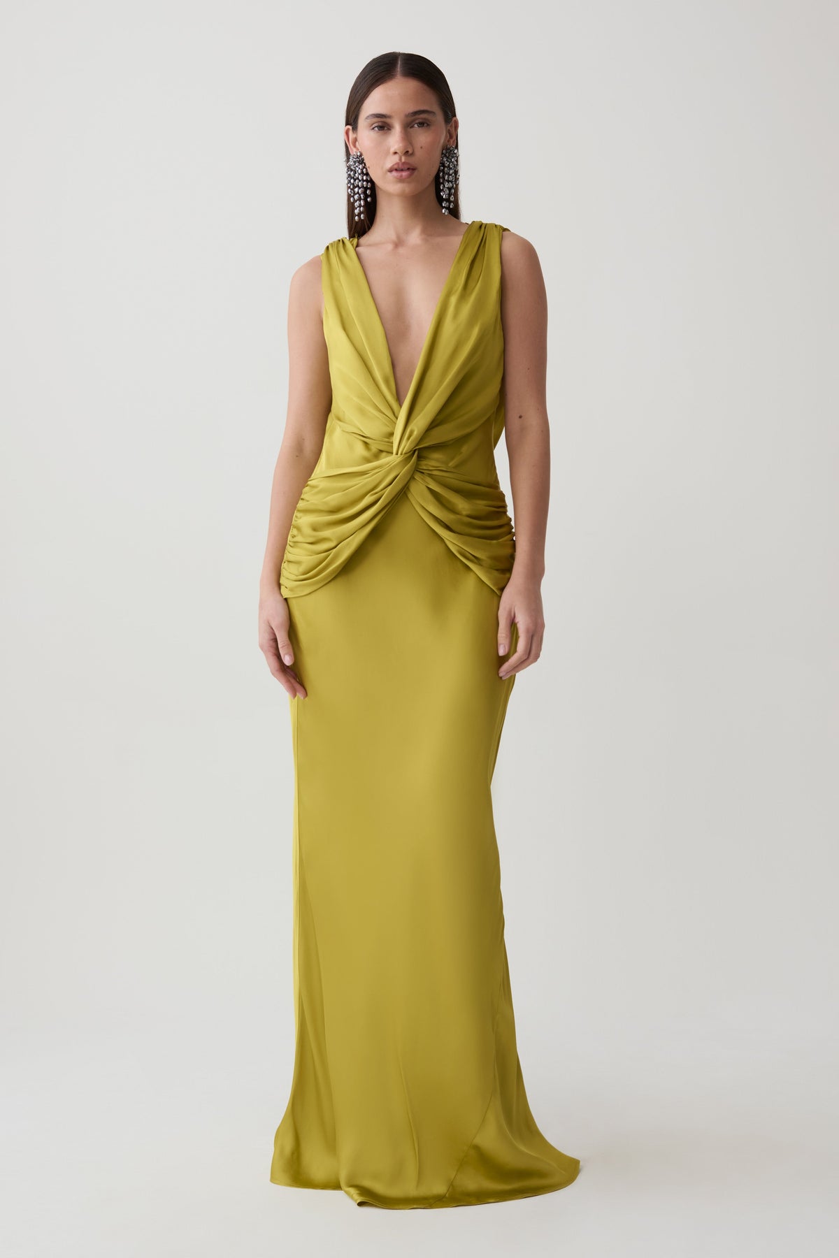 A woman modeling a sleeveless, floor-length olive yellow gown with a deep V-neckline and a twisted knot detail at the waist, set against a plain light gray studio background.