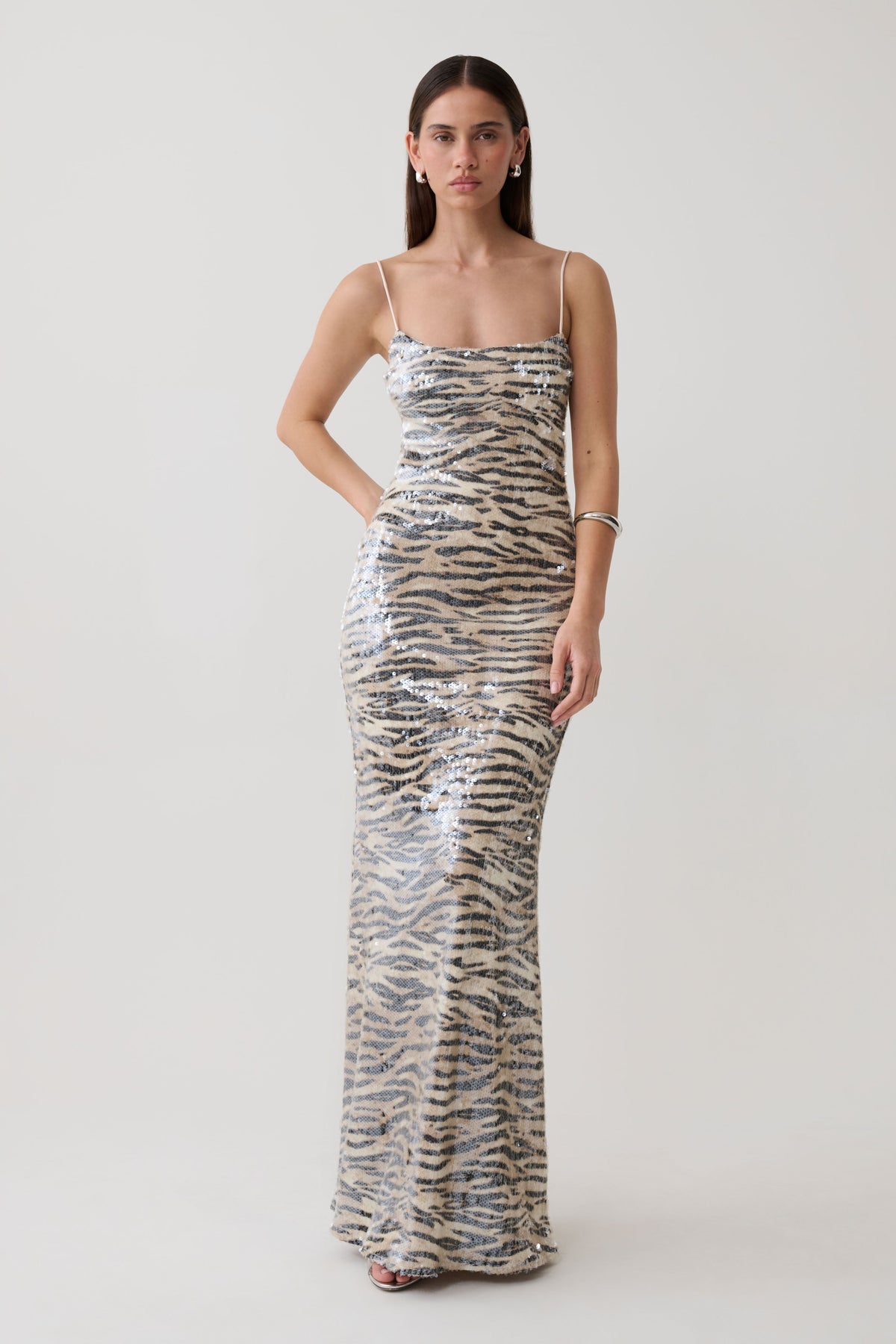 A woman modeling a floor-length, spaghetti-strap sequin gown with a beige, silver, and black animal-stripe pattern against a plain light gray studio background.