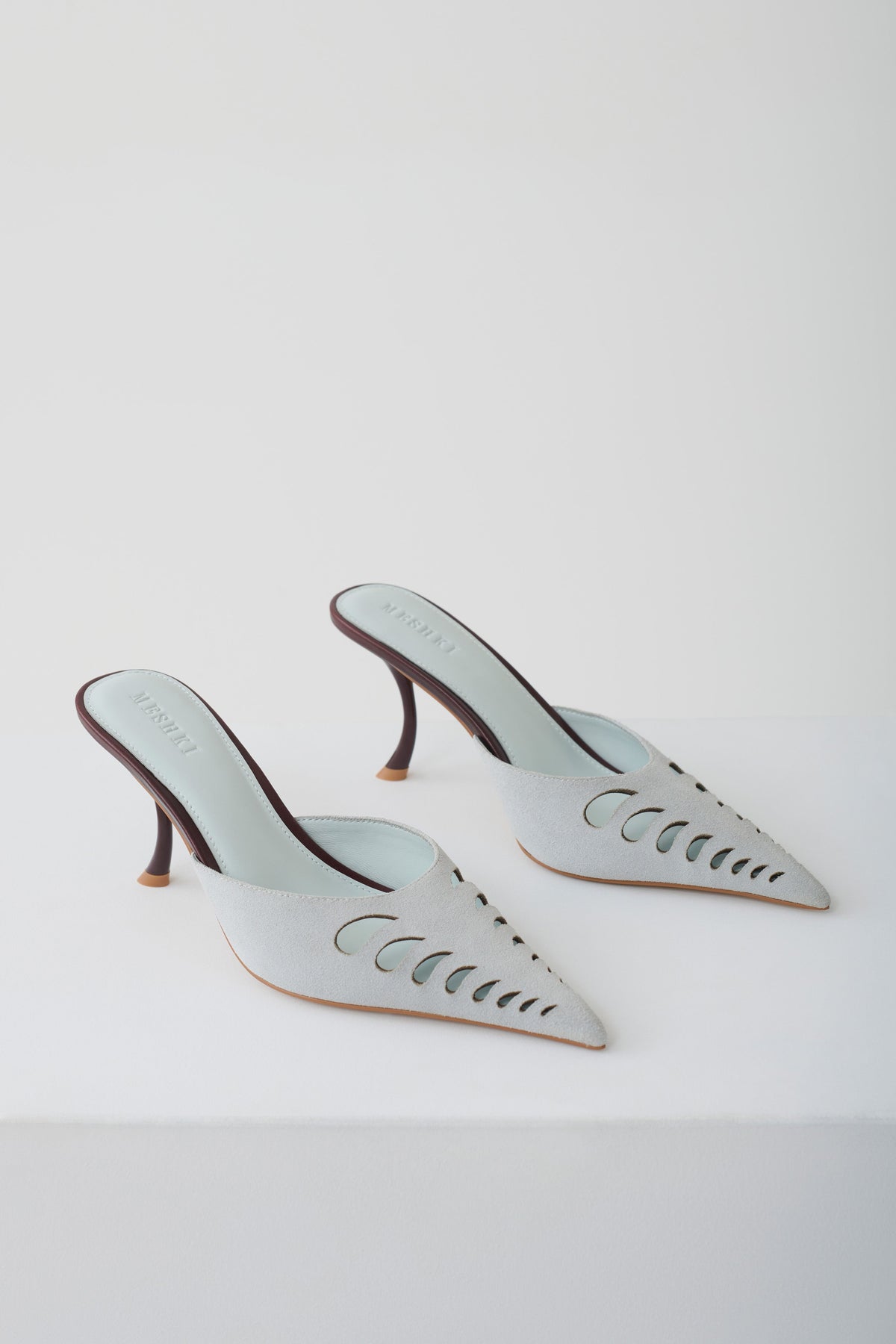 A pair of pointed-toe mule heels in a pale icy blue color, featuring decorative cutout detailing on the sides and slim dark brown stiletto heels, photographed on a clean white studio background.