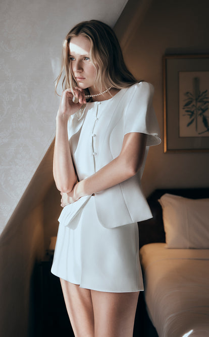 Woman in a white outfit standing in a room with a bed and framed picture in the background.