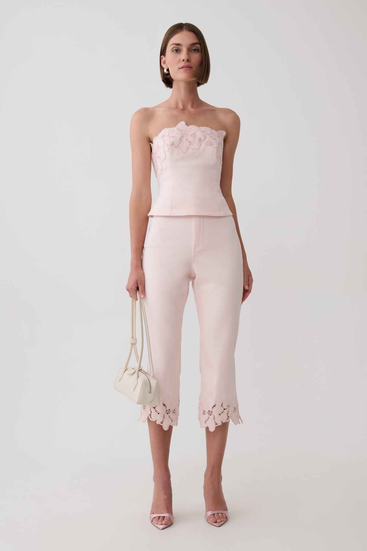 A model stands against a light gray studio background wearing a pale pink strapless top with ruffled detailing and matching cropped pants with floral lace trim at the hems. She is holding a small cream handbag and wearing clear high-heel sandals.