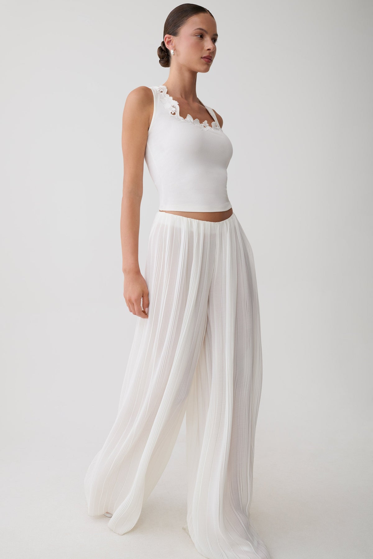 A woman modeling a white sleeveless crop top with scalloped trim and matching white wide-leg pleated pants against a plain light gray studio background.