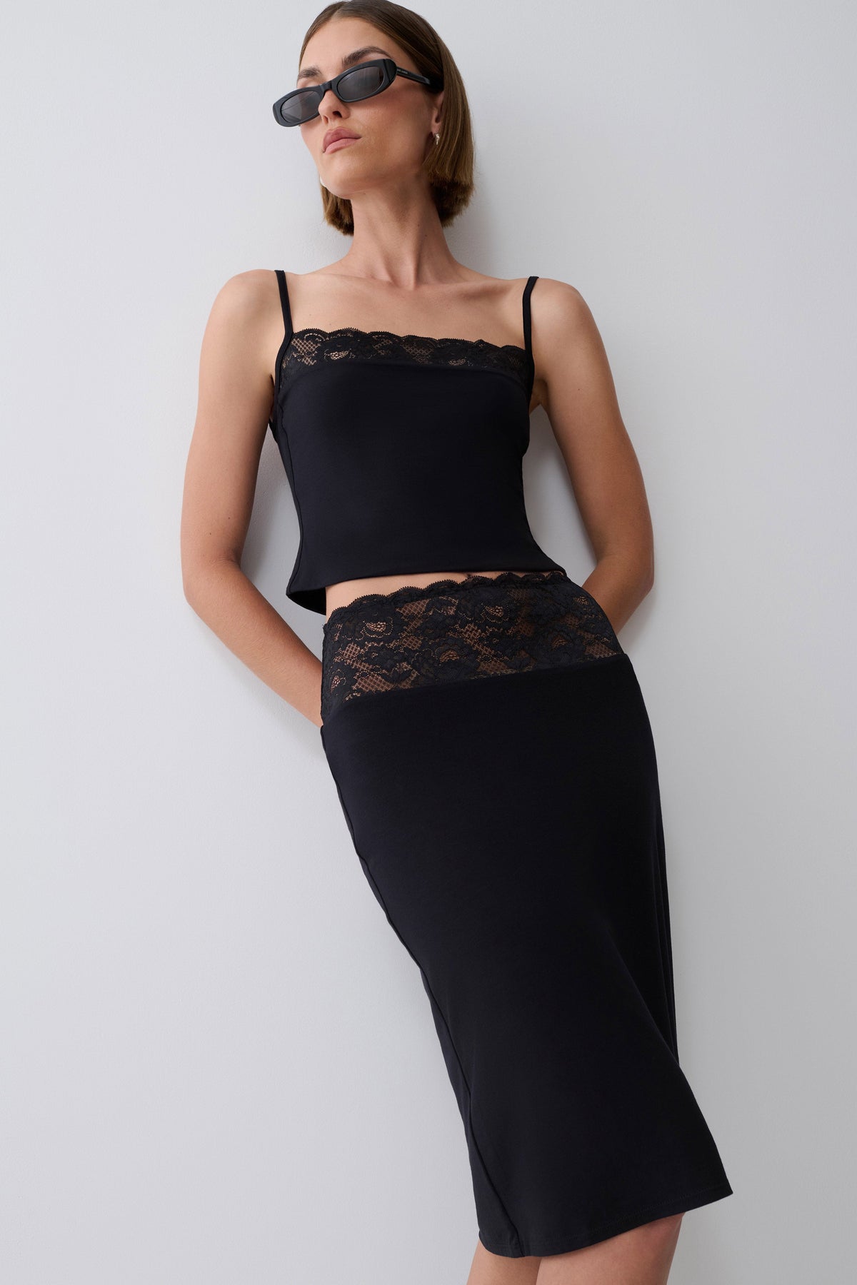 A woman modeling a black two-piece set consisting of a spaghetti-strap crop top with lace trim and a matching high-waisted pencil skirt with lace detailing, styled with dark sunglasses against a plain light gray wall.