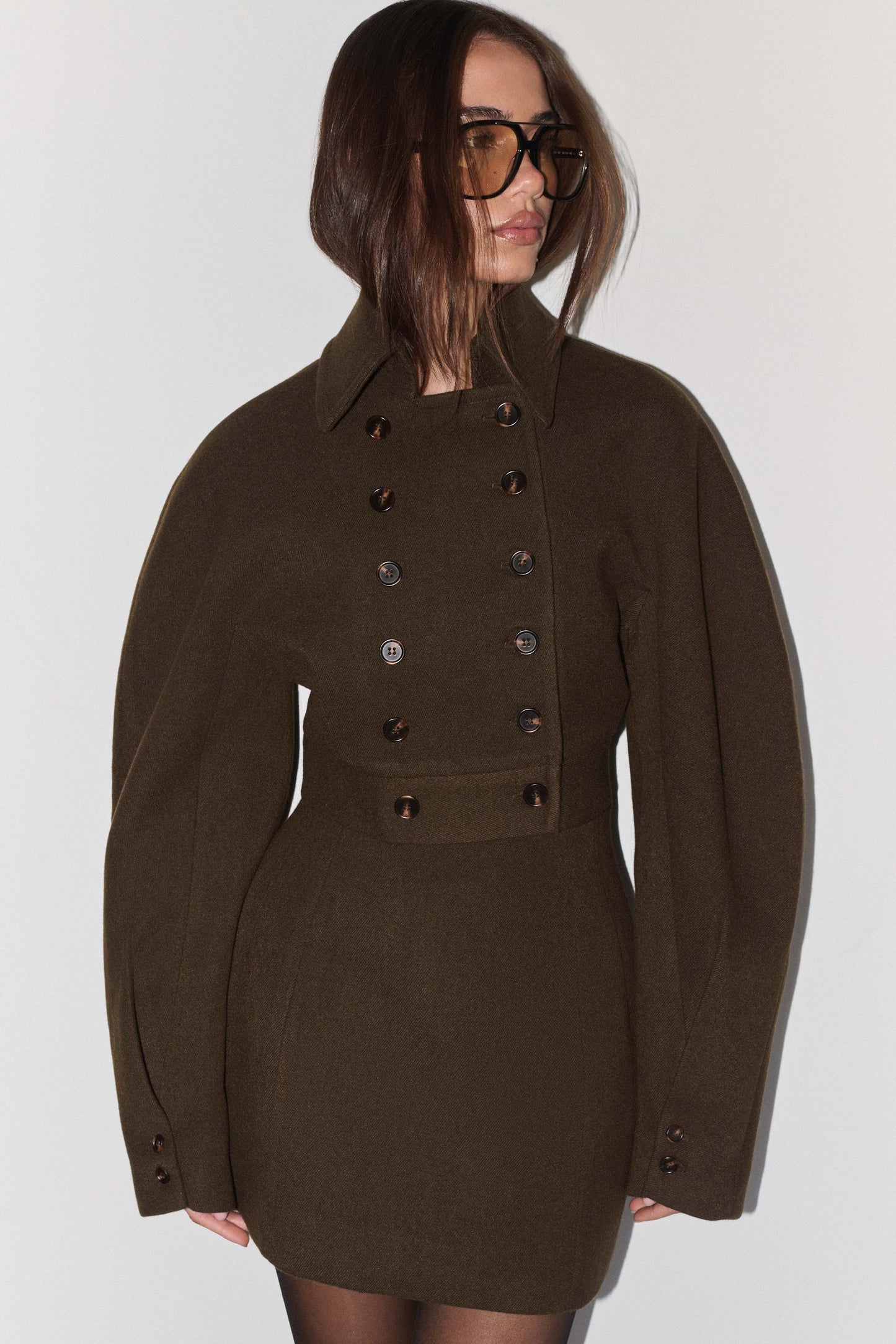 A woman wearing a dark olive brown double-breasted jacket with oversized long sleeves and dark brown buttons, paired with matching mini skirt, black tights and glasses, against a plain light gray background.