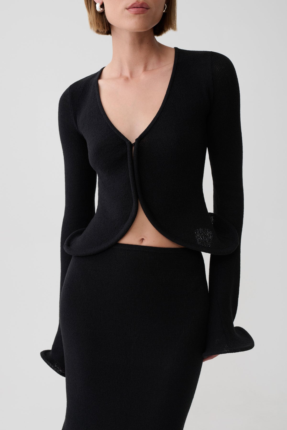 A model wearing a black long-sleeve knit cardigan with a deep V-neck and flared cuffs, paired with a matching black skirt, against a light gray studio background.
