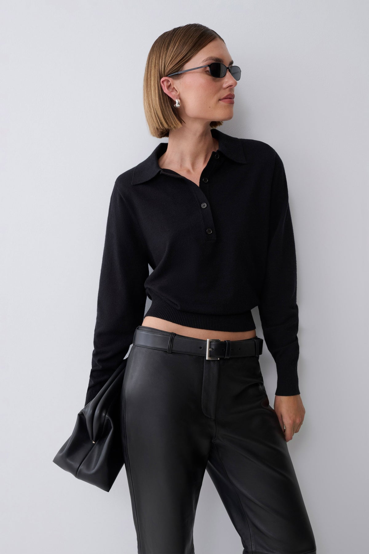 A woman wearing a black long-sleeve cropped polo shirt, black leather-look pants, a black belt, and sunglasses, holding a black clutch against a plain light gray background.