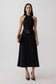 A woman modeling a sleeveless black midi dress with a halter-style neckline, a matching black belt with a gold buckle, black pointed-toe heels, and a small black clutch, photographed against a light gray studio background.
