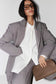 A model wearing a gray tailored blazer and matching trousers over a white button-up shirt, styled with oversized black sunglasses and a brown leather clutch against a light gray studio background.