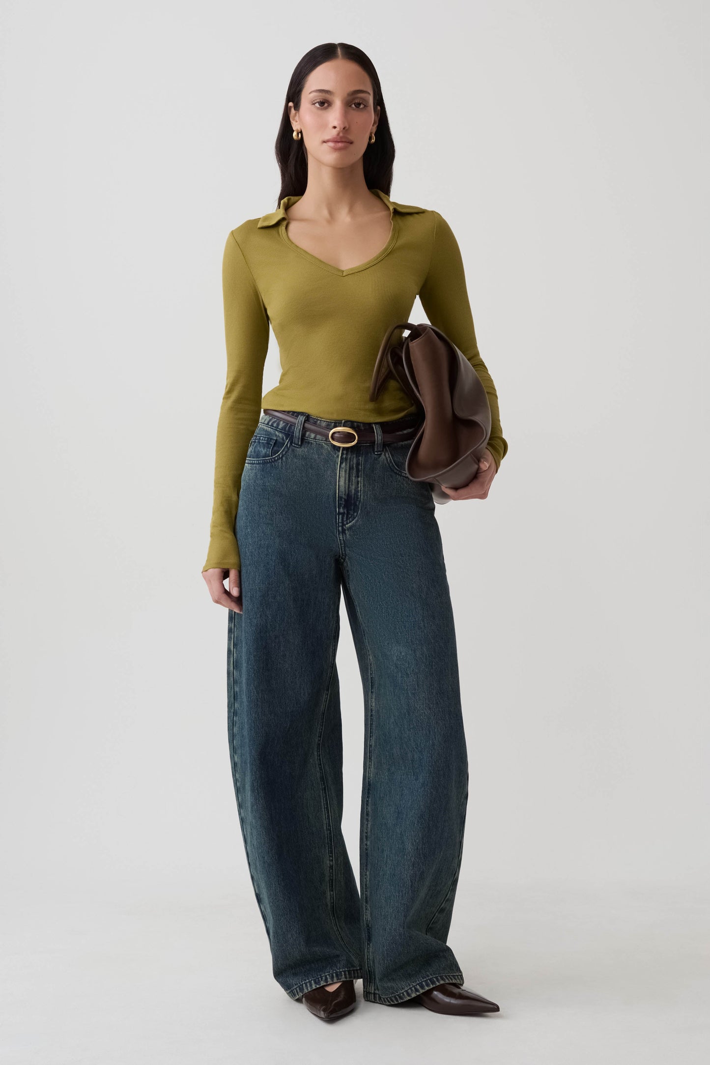 A woman stands against a light gray studio background wearing a fitted olive green long-sleeve top, wide-leg dark blue jeans with a brown belt, dark brown pointed flats, and holding a brown leather clutch.