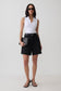 A model stands against a light gray studio background wearing a white sleeveless top, black high-waisted shorts with a black belt, black sandals, and holding a black clutch.