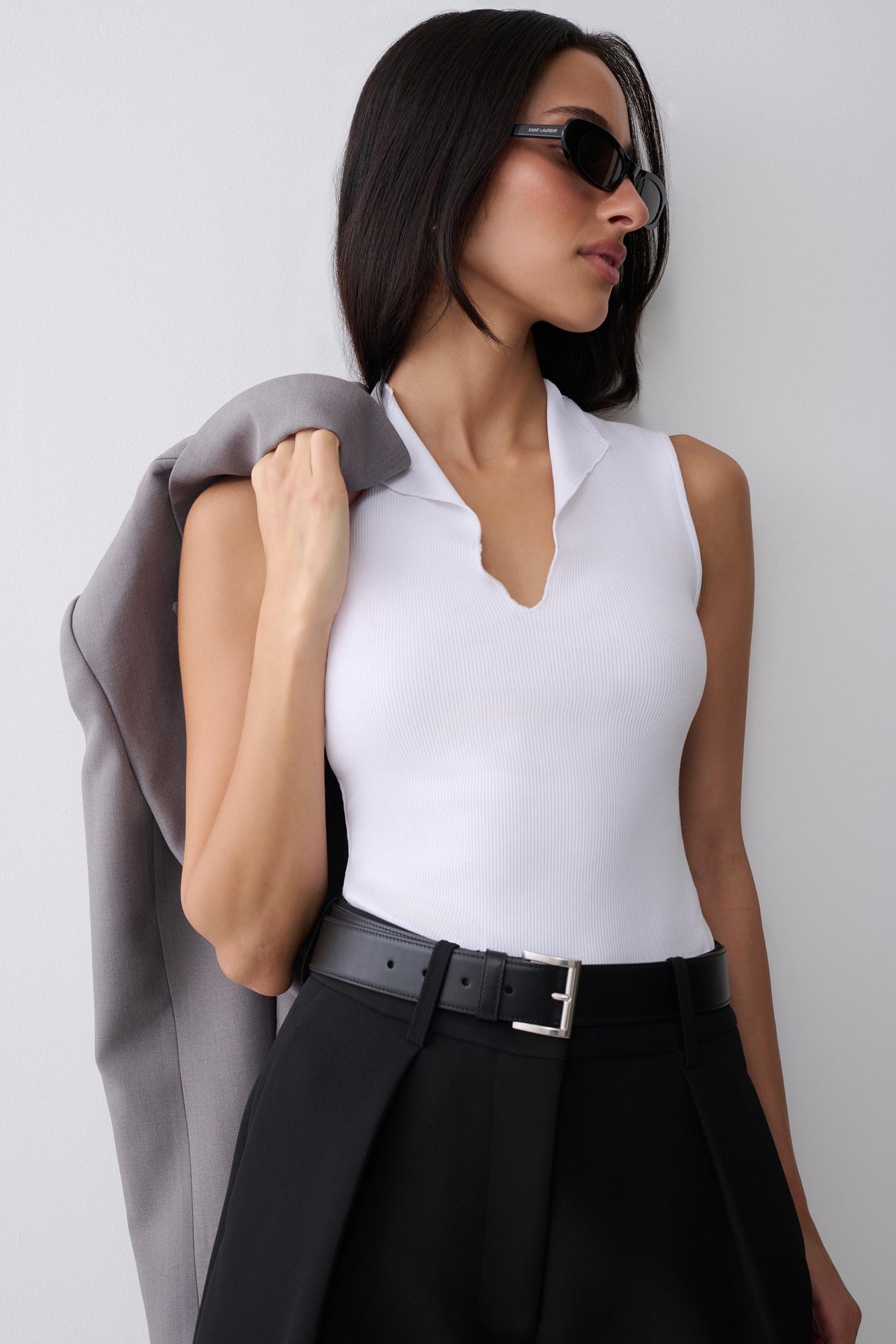 A woman wearing a fitted white sleeveless top with a split neckline, styled with black high-waisted trousers, a black belt, and dark sunglasses, while holding a gray blazer against her shoulder against a light gray background.