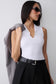 A woman wearing a fitted white sleeveless top with a split neckline, styled with black high-waisted trousers, a black belt, and dark sunglasses, while holding a gray blazer against her shoulder against a light gray background.