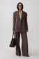 A model wearing a brown double-breasted pinstripe suit with matching wide-leg trousers, a white collared shirt, glasses, and holding a brown leather handbag against a light gray studio background.
