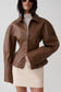 A woman wearing a cropped brown faux-leather jacket with a front zipper, collar, and flared sleeves, paired with a cream mini skirt against a light gray studio background.