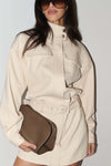 A woman wearing a cream-colored denim mini dress with a belt and a brown leather clutch bag, set against a plain light gray background.