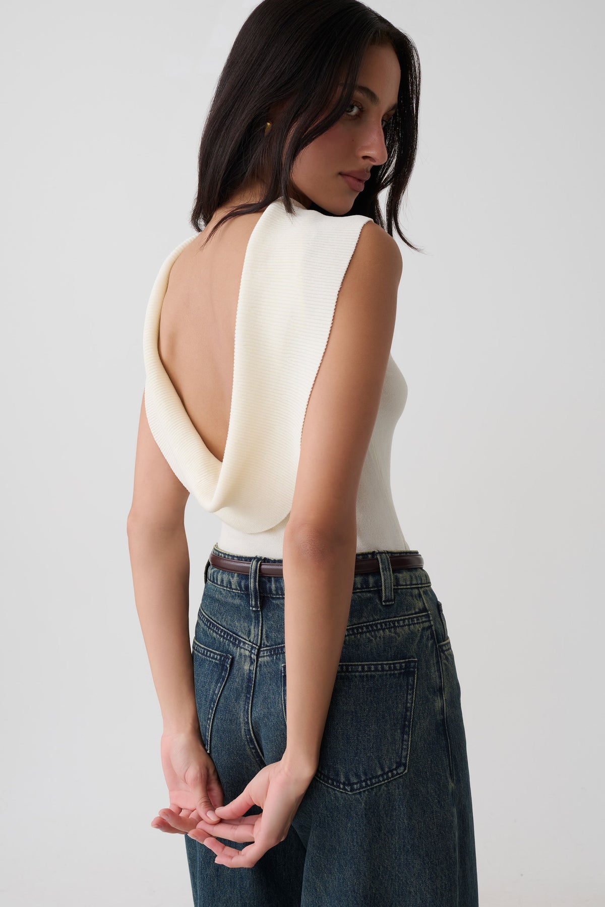 A woman wearing a cream-colored sleeveless top with a dramatic draped open back, paired with high-waisted dark blue jeans, photographed against a plain light gray studio background.