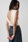 A woman wearing a cream-colored sleeveless top with a dramatic draped open back, paired with high-waisted dark blue jeans, photographed against a plain light gray studio background.