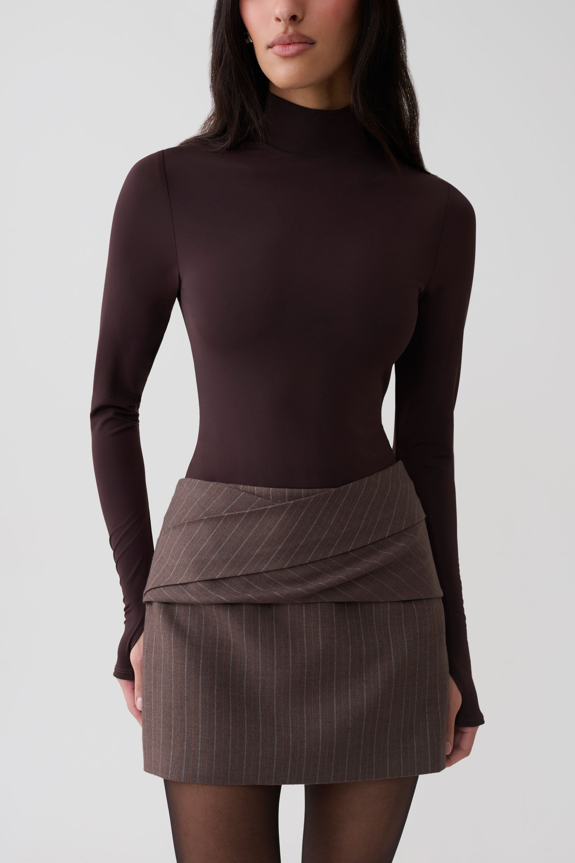 A model wearing a dark brown mock neck long-sleeve top and a brown pinstriped wrap mini skirt, photographed against a light gray studio background.