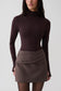 A model wearing a dark brown mock neck long-sleeve top and a brown pinstriped wrap mini skirt, photographed against a light gray studio background.