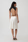 A woman shown from the back wearing a sleeveless black top with an open back and a white knee-length skirt, photographed against a plain light gray studio background.