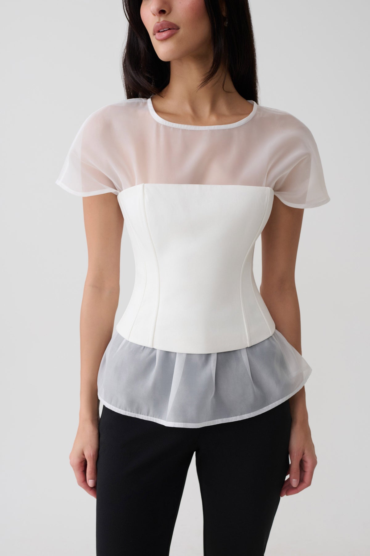 A woman wearing a white corset-style short-sleeve top with sheer mesh overlay panels on the shoulders and hem, paired with black pants, against a light gray studio background.