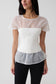 A woman wearing a white corset-style short-sleeve top with sheer mesh overlay panels on the shoulders and hem, paired with black pants, against a light gray studio background.
