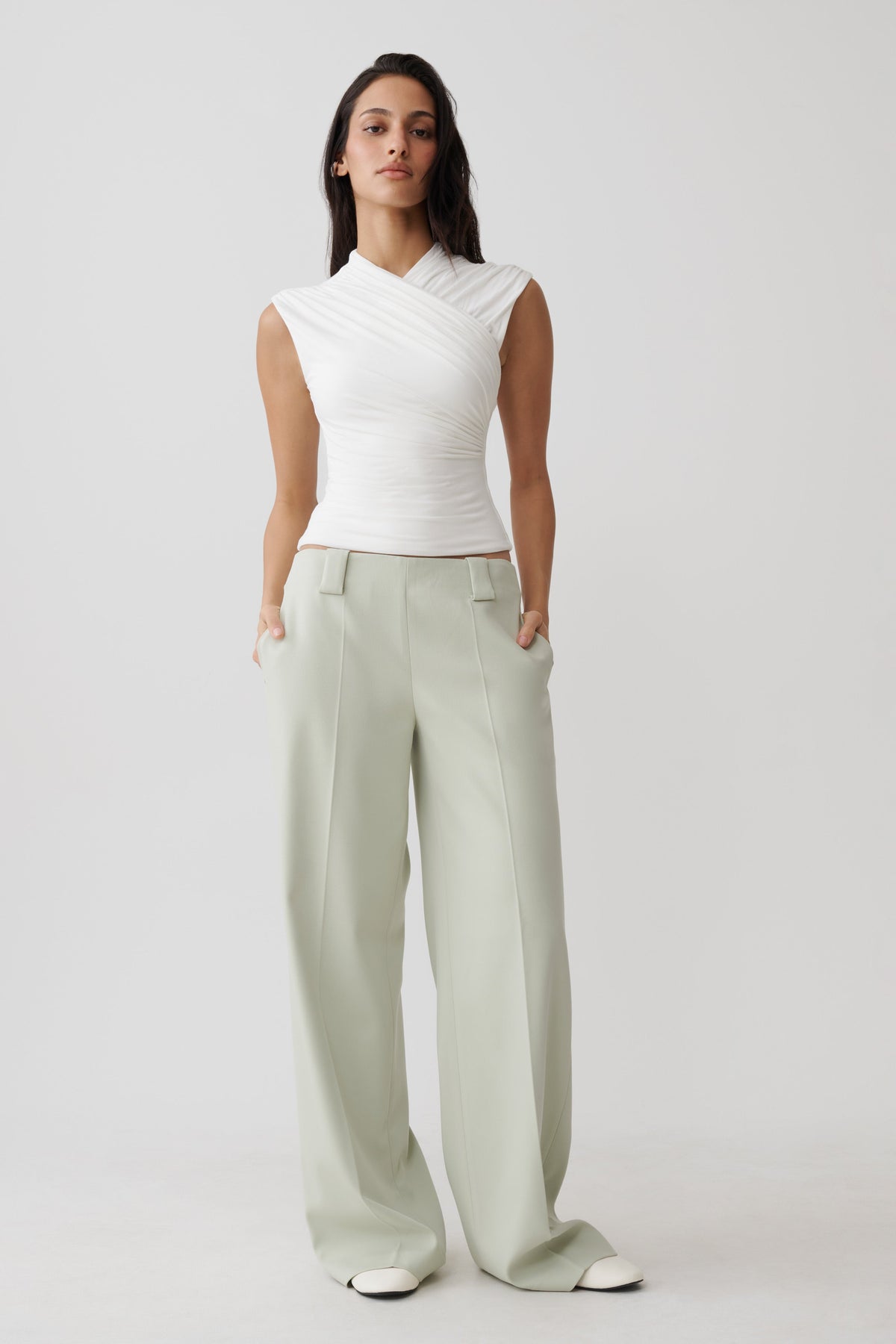 A woman modeling a white sleeveless wrap-style top and light sage green wide-leg trousers against a plain light gray studio background.