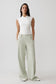 A woman modeling a white sleeveless wrap-style top and light sage green wide-leg trousers against a plain light gray studio background.