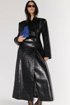 A model wearing a long, structured black faux-leather coat with a glossy, embossed texture, holding a blue velvet clutch against a plain light gray studio background.