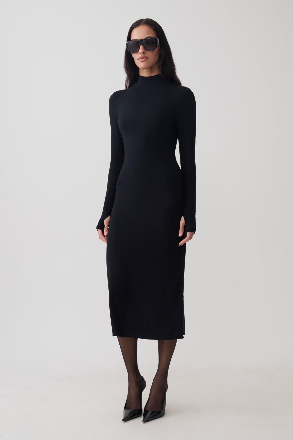 A woman modeling a fitted black long-sleeve mock-neck midi dress, styled with black sunglasses, sheer black tights, and black pointed-toe heels against a plain light gray studio background.