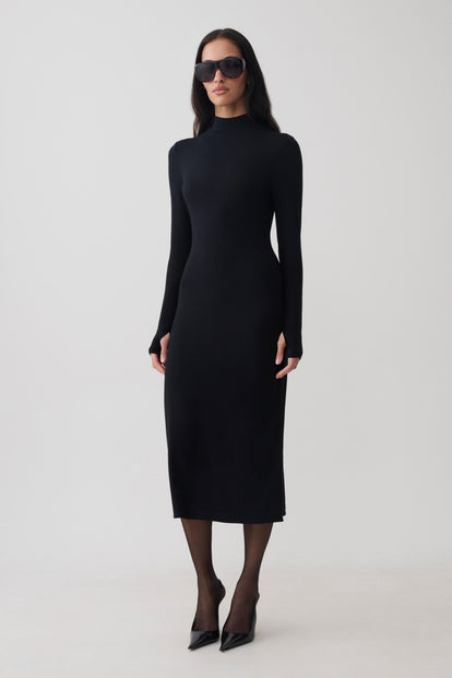 A woman modeling a fitted black long-sleeve mock-neck midi dress, styled with black sunglasses, sheer black tights, and black pointed-toe heels against a plain light gray studio background.