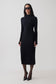 A woman modeling a fitted black long-sleeve mock-neck midi dress, styled with black sunglasses, sheer black tights, and black pointed-toe heels against a plain light gray studio background.