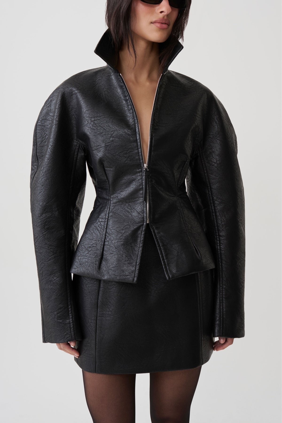 A model wearing a black textured faux-leather jacket with a high collar and a matching black mini skirt, photographed against a light gray studio background.