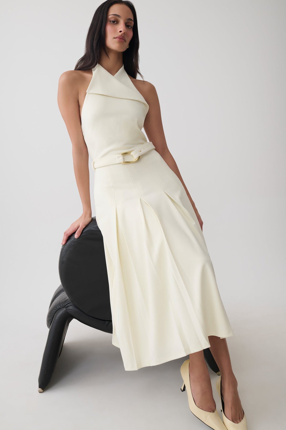 A model wearing a cream-colored halter midi dress with a matching belt, posing on a black sculptural chair against a light gray studio background.