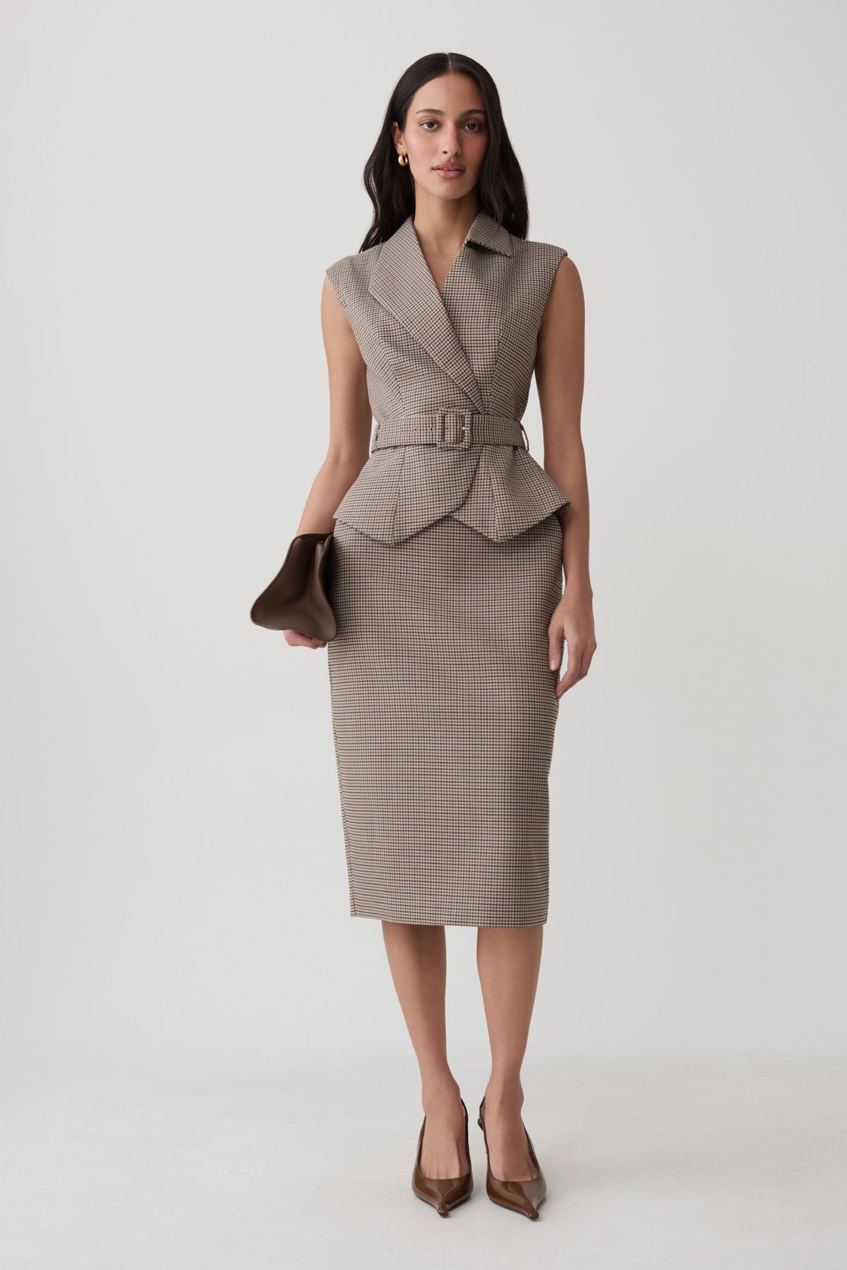 A woman models a sleeveless taupe houndstooth vest and matching pencil skirt set, holding a brown clutch against a light gray studio background.