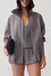 A woman models an oversized gray zip-up jacket with a stand collar, paired with matching shorts and a matching camisole. She holds a small black clutch against a clean, light gray studio background.