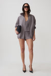 A model stands against a light gray studio background wearing a coordinated gray outfit consisting of an oversized zip-front jacket, matching top, and shorts. She is also wearing black pointed heels and holding a small dark clutch.