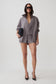 A model stands against a light gray studio background wearing a coordinated gray outfit consisting of an oversized zip-front jacket, matching top, and shorts. She is also wearing black pointed heels and holding a small dark clutch.