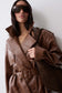 A woman wearing a brown leather trench coat with a matching belt and large dark sunglasses, holding a dark brown shoulder bag against a plain light gray background.