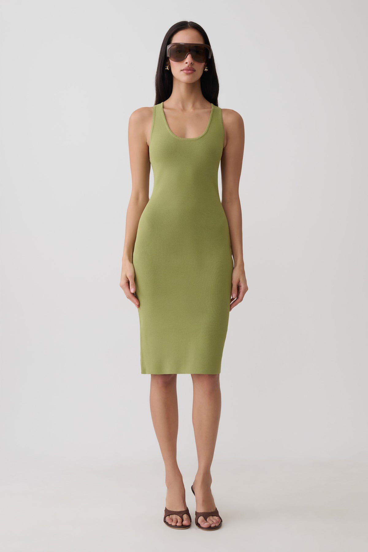 A woman modeling a sleeveless olive green bodycon midi dress with a scoop neckline, paired with dark sunglasses and brown strappy heels, against a plain light gray studio background.