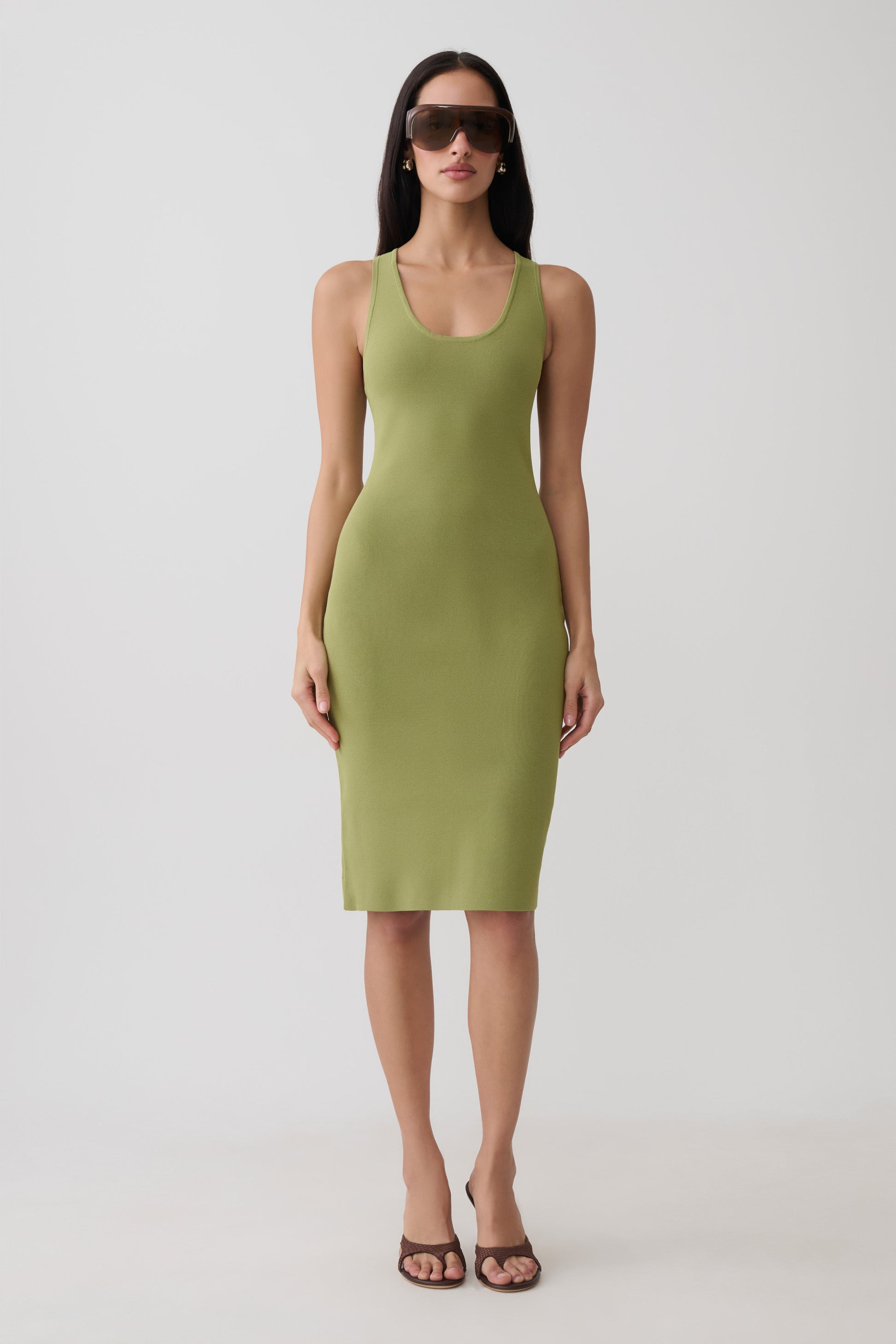 A woman modeling a sleeveless olive green bodycon midi dress with a scoop neckline, paired with dark sunglasses and brown strappy heels, against a plain light gray studio background.
