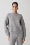 Woman wearing a gray sweater and pants on a plain background.