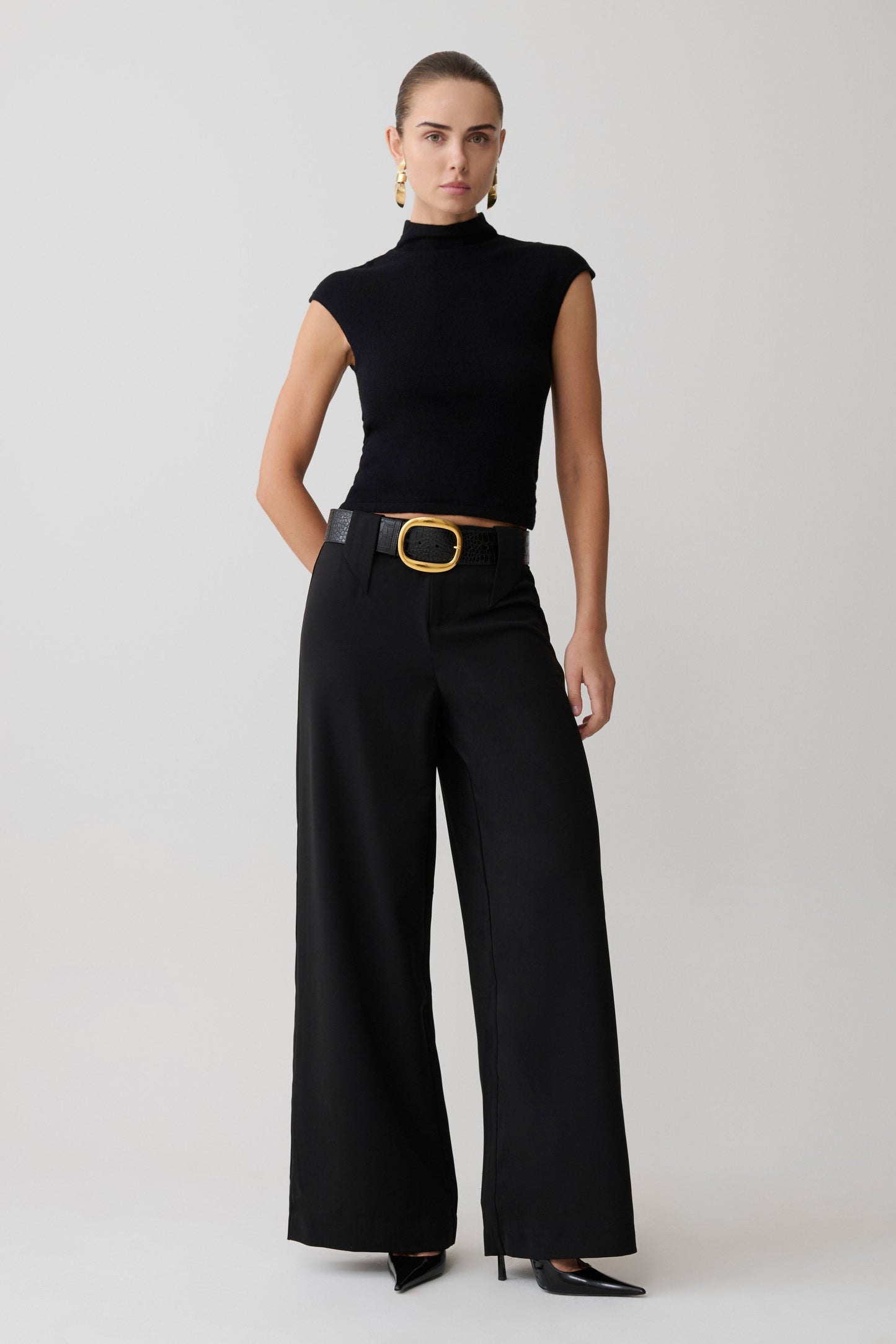 Woman wearing a black sleeveless top and wide-leg pants with a gold belt on a white background