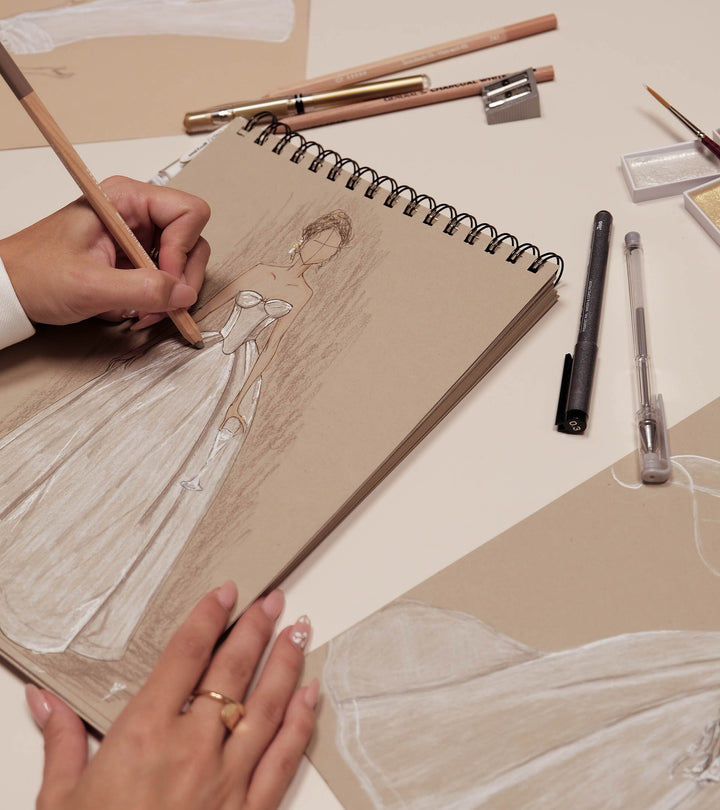 Person drawing a figure in a sketchbook with various art supplies on a table.